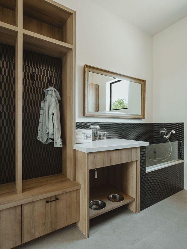 A contemporary bathroom with a built-in timber vanity and open shelving, integrated pet-bowl storage, a large framed mirror, and a bathtub clad in dark patterned tile. The room combines pale timber joinery, a light benchtop, white walls, and a grey floor with a practical, refined aesthetic.