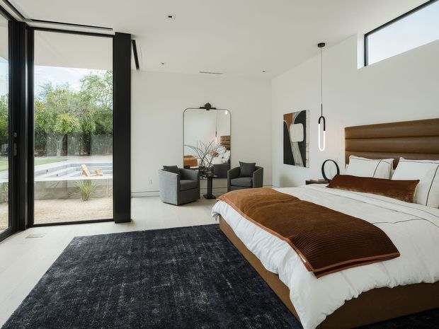 Bright contemporary bedroom with a large upholstered bed, white bedding, brown throw and cushions, dark area rug, two armchairs, full-height black-framed glazing, and a minimalist pendant light. A pool and landscaped garden are visible outside through the glazing.