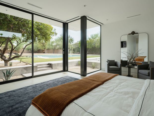 Bright modern bedroom with a low-profile bed dressed in white bedding and a rust-brown throw, dark charcoal rug, and two dark upholstered armchairs. Full-height black-framed glazing wraps the corner, opening views toward a landscaped outdoor area and swimming pool. A minimalist console and rounded-edge mirror sit against the white wall, with a dark structural column defining the room.