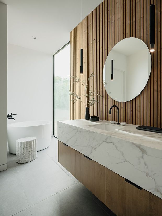A contemporary bathroom featuring a long double vanity with a thick white veined stone benchtop, timber cabinetry, and a vertical timber-slat feature wall. Two black pendant lights hang above the vanity, complemented by a large round mirror and black tapware. A freestanding white bathtub sits beside full-height glazing, with pale large-format floor tiles and a sculptural white stool creating a bright, refined atmosphere.