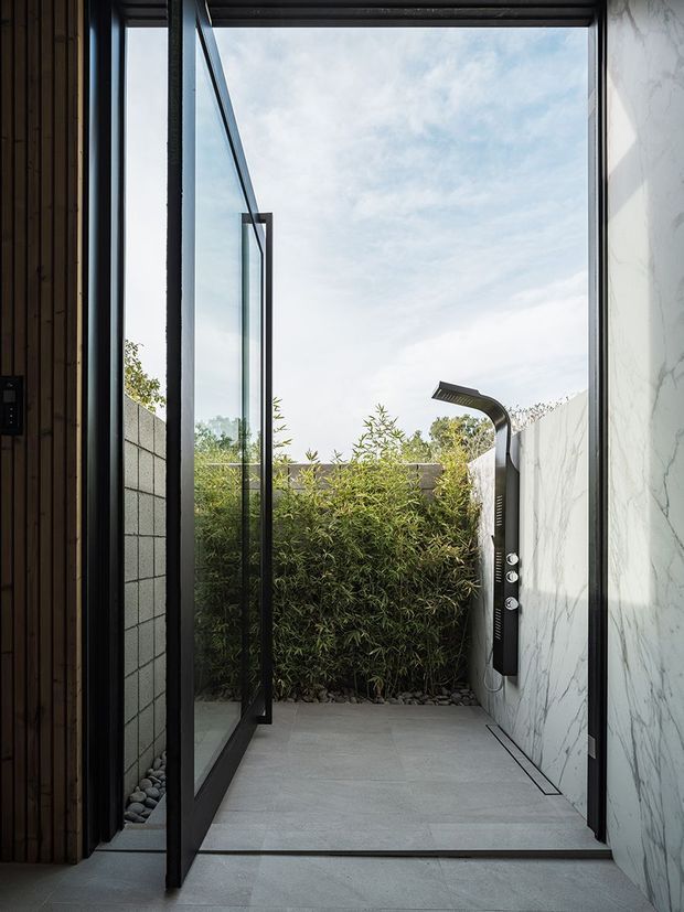 A narrow, modern outdoor shower courtyard viewed from an interior threshold. The space features large-format grey floor tiles, a dramatic white marble-look wall, black-framed full-height glazing and a frameless glass shower door, with a dark sculptural shower fixture and dense bamboo screening at the far end. Vertical timber cladding is visible along the left edge.
