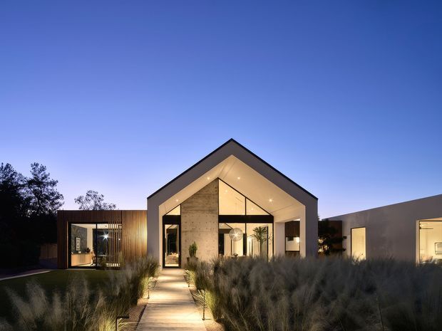 Twilight exterior view of a contemporary single-storey residence with a prominent gabled central volume, large glazed entry façade, warm recessed lighting, timber-clad side wing, and low flat-roofed wing. A paved path leads through landscaped ornamental grasses toward the entrance.