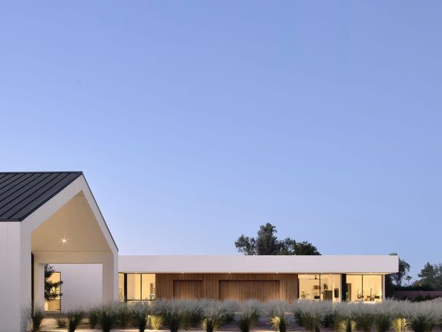 Low, contemporary single-storey residence viewed from the landscaped front yard at dusk. The composition features a prominent white gabled entrance volume with dark standing-seam metal roofing, connected to a long flat-roofed wing with extensive glazing and warm timber-clad walls. Ornamental grasses and drought-tolerant planting occupy the foreground.