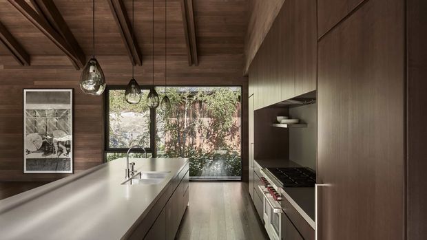 A narrow, refined contemporary kitchen viewed along a long island toward a large garden-facing window. The room features dark timber wall and ceiling lining with exposed ceiling beams, full-height dark timber cabinetry, a pale stone island benchtop with integrated sink and tapware, a built-in cooktop and oven bank, open shelving, pendant lights, and timber flooring. The palette is warm and restrained, with strong natural light and greenery visible beyond the glazing.