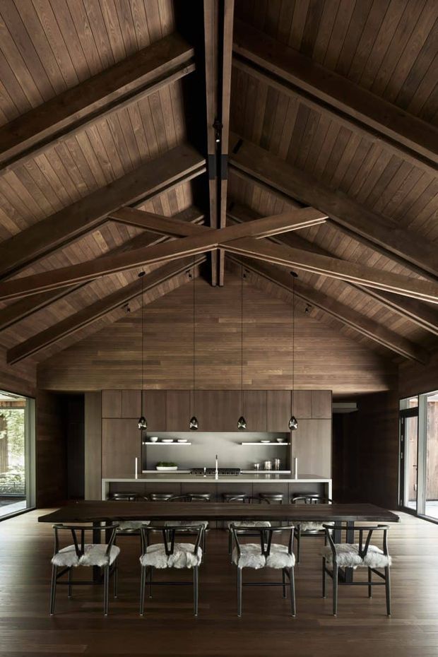 A dramatic open-plan dining and kitchen interior beneath a tall exposed timber gable roof. Dark wood panelling covers the walls, ceiling and cabinetry, with prominent exposed timber beams and rafters. A long dark dining table with multiple black-framed chairs occupies the foreground, while a linear kitchen with an island, stools, pendant lights and integrated cabinetry sits behind it. Large glazed openings bring in natural light from both sides.
