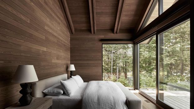 A compact, warm bedroom lined in dark timber, with a bed positioned beside expansive floor-to-ceiling corner glazing overlooking a wooded setting. The room features an exposed timber ceiling with beams, dark timber wall panelling, a neutral upholstered bed, bedside table and lamps, and a strong connection to the surrounding forest.