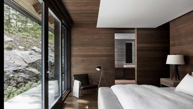 A modern bedroom with a large bed, dark timber wall panelling, timber flooring, and expansive floor-to-ceiling glazing opening toward a rocky natural landscape. A compact seating area and floor lamp sit beside the window, while a bathroom or dressing area is visible through the opening at the rear.