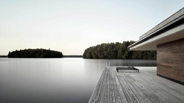 A minimalist lakeside house viewed from a broad timber deck extending over calm water. The low, flat-roofed structure has horizontal timber wall cladding, a projecting roof edge, and a simple dark metal balustrade. Two lounge chairs sit on the deck, with wooded islands or shoreline across the lake creating a quiet, secluded setting.