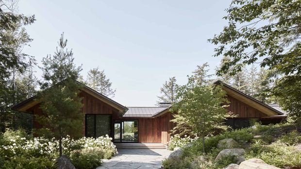 Single-storey residential building set within a landscaped garden, composed of several connected timber-clad wings with pitched gabled roofs. Large dark-framed glazed openings and a central paved entry path are visible, with mature planting, rocks and shrubs surrounding the structure.