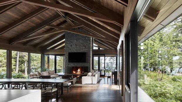 A spacious, open-plan pavilion-like living and dining area with extensive floor-to-ceiling glazing overlooking a wooded landscape. The room features a high pitched timber ceiling with exposed rafters and beams, dark timber flooring and structural framing, a central dark masonry fireplace with a lit fire, a dining table and chairs in the foreground, and a seating area beyond. The atmosphere is warm, natural, and strongly connected to the outdoors.