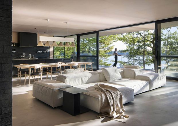 Open-plan contemporary living and dining area with a sleek black kitchen and long island, pale timber dining table and chairs, and a large low-profile cream sectional sofa. Full-height black-framed glazing opens the room to a leafy waterfront or lakeside outlook. The space has a polished, understated aesthetic with generous natural light and strong indoor-outdoor connection.