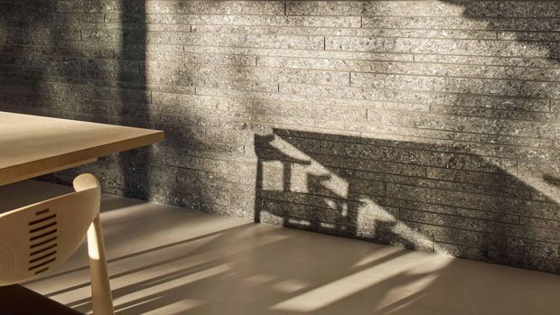 A minimalist interior corner with a pale timber dining table and chair beside a textured horizontally coursed grey masonry wall. Strong low-angle sunlight casts dramatic geometric shadows across the wall and smooth floor, creating a warm, quiet atmosphere.