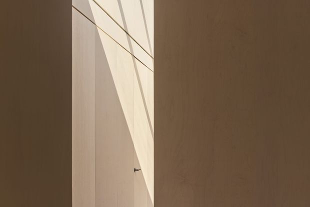Minimal architectural interior detail showing a narrow vertical opening or passage between warm brown timber-clad wall planes. Strong sunlight enters from above or the side, creating angular geometric shadows and a bright cream-toned illuminated surface. A small dark door handle is visible in the recess. The composition is abstract, quiet, and highly minimalist, with no furnishings or clear room function visible.