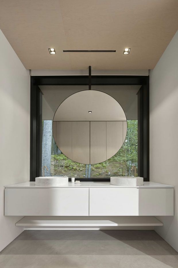 A minimalist contemporary bathroom vanity with a floating white double cabinet, two round vessel basins, wall-mounted taps, and a large circular suspended mirror. The room has pale smooth walls and ceiling, a dark-framed full-height window looking onto trees, recessed ceiling lights, and a large-format grey floor.