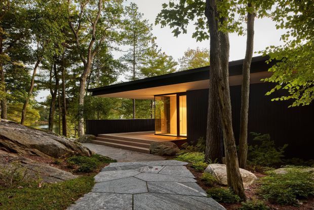 A low, dark-clad modern house nestled among mature trees and exposed rock, approached by a broad irregular stone-paved path. The building features a dramatically projecting flat roof, extensive warm-lit glazing, a timber-toned entry, dark vertical screening, and a raised timber deck with steps. The wooded setting and integrated landscape give the image a secluded, nature-connected character.