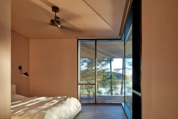 A compact, warm-toned bedroom with pale timber wall and ceiling linings, a low bed, ceiling fan, and a large floor-to-ceiling glazed corner opening onto a timber deck and leafy landscape. Dark slim-framed glazing and built-in joinery create a simple contemporary retreat with strong indoor-outdoor connection.