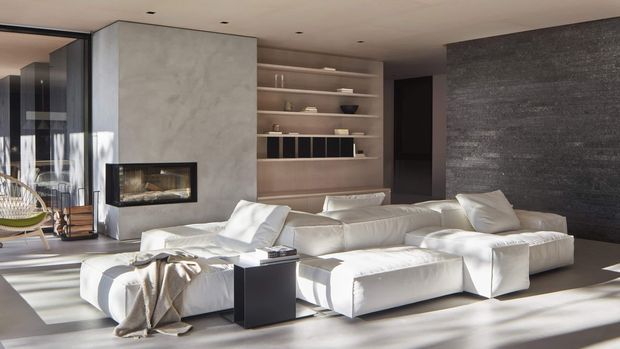 A spacious contemporary living room with a low white sectional sofa arranged around a dark square side table and a built-in linear fireplace. The room features a large pale concrete or microcement fireplace surround, warm timber open shelving and cabinetry, a dark textured feature wall, recessed ceiling lights, and expansive glazing with strong sunlight and views into an adjacent outdoor area. The palette is neutral, warm, and refined.