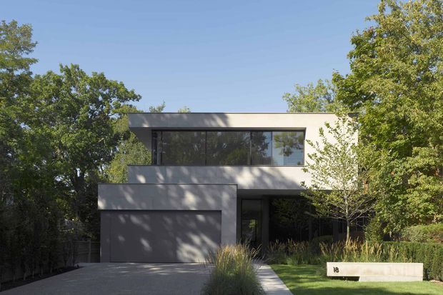 A modern two-storey residential house viewed from the front, partially screened by mature trees and landscaped planting. The building has a strong rectilinear composition, a flat roof, pale rendered or concrete-like walls, expansive dark-framed upper-level glazing, and an integrated single garage. A paved driveway and path lead through a restrained garden with ornamental grasses and clipped hedges.