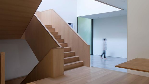 A minimalist contemporary interior featuring a sculptural timber staircase with enclosed side panels and integrated handrails. The staircase rises from a light timber floor toward an upper level, opening into a bright double-height circulation space with white walls, pale flooring, and a large rectangular ceiling opening. A simple timber bench or projecting surface is visible at right, while a blurred person provides a sense of scale.