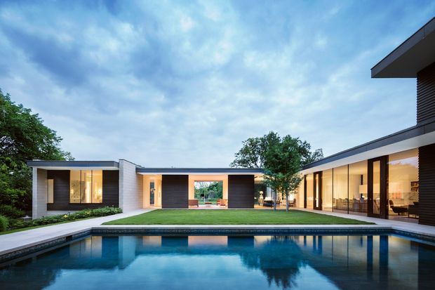 A low-slung contemporary residence arranged around a landscaped courtyard and rectangular swimming pool. The composition features flat rooflines, dark horizontal exterior cladding, crisp white rendered or masonry planes, extensive floor-to-ceiling glazing, and warm interior lighting visible through the windows. Mature trees frame the property, while the pool reflects the house and evening-blue sky.