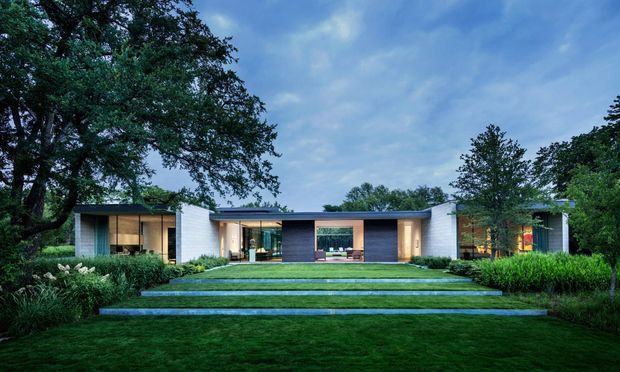 A wide exterior view of a low, single-storey modern residence set within a landscaped garden. The house has a long, horizontally composed flat roof, white masonry or rendered volumes, dark vertical cladding at the central entry, extensive floor-to-ceiling glazing, and a broad stepped lawn leading to the facade. Mature trees and dense planting frame the property.