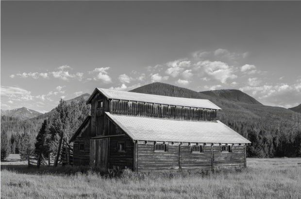 Black-and-white photograph of a rustic timber barn or mountain lodge set in an open meadow, with a steeply pitched corrugated roof, weathered horizontal timber siding, a raised upper-level hayloft or clerestory section, and surrounding forested mountains.