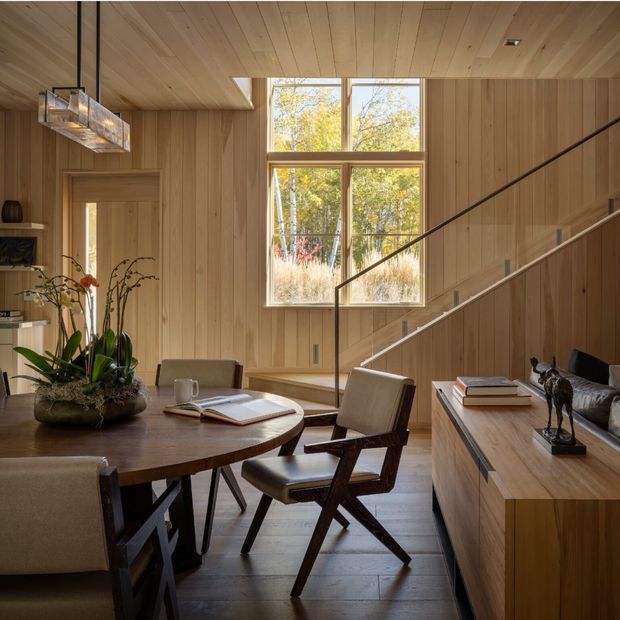 Warm, wood-lined open-plan interior combining a dining area and living space beneath a timber-clad ceiling. A round dark wood dining table with upholstered chairs occupies the foreground, while a built-in timber cabinet and sofa sit to the right. A sculptural pendant light, large timber-framed window, indoor plants, books and artwork add detail. A stair with slim dark metal railing rises along the right wall, and the room is filled with natural light and views of trees.