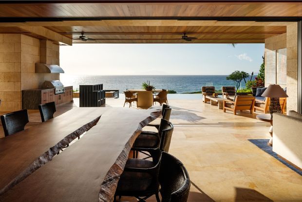Open-plan coastal entertaining space with a large live-edge dining table, black dining chairs, an outdoor kitchen and barbecue to the left, and a separate outdoor dining and lounge area opening directly toward the ocean. The space features extensive timber ceiling lining and beams, warm stone-clad walls, large open apertures, and pale stone flooring, creating a luxurious indoor-outdoor connection.