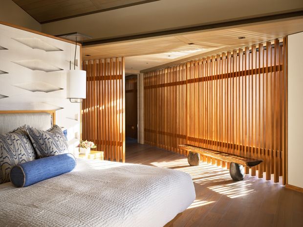 Warm, sunlit bedroom with a low platform bed, textured white bedding, patterned pillows, and a blue bolster. A dramatic timber slatted screen defines the hallway and creates strong linear shadows across the timber floor. Built-in white shelving and pendant lighting contribute to the refined mid-century-inspired interior.