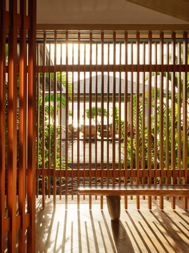 A warm, sunlit transitional space framed by a dense timber screen of vertical slats. The view looks toward a leafy internal courtyard with a tiled floor, outdoor seating, and abundant tropical planting. Timber framing, slatted screening, a low timber bench, and strong linear shadows create a tranquil, indoor–outdoor atmosphere.