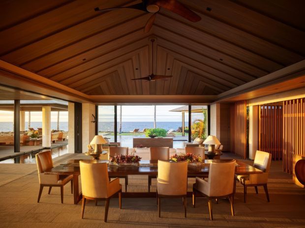 A warm, sophisticated dining room with a long timber dining table and upholstered dining chairs, opening through extensive glazing to a beachfront pool and ocean view. The room features a richly detailed timber-lined vaulted ceiling with exposed geometric framing, timber flooring, recessed lighting, a sculptural ceiling fan, and a vertical timber slat feature wall. Table lamps and floral arrangements add a refined resort-like atmosphere.