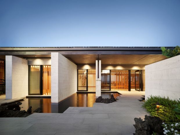 Contemporary single-storey courtyard-style residence with a low flat roof, pale masonry walls, warm timber soffit, extensive vertical timber screening, dark-framed glazing, and broad grey stone paving. A shallow reflecting pool and sculptural dark rocks create a refined entry courtyard, with planted greenery at the edges.
