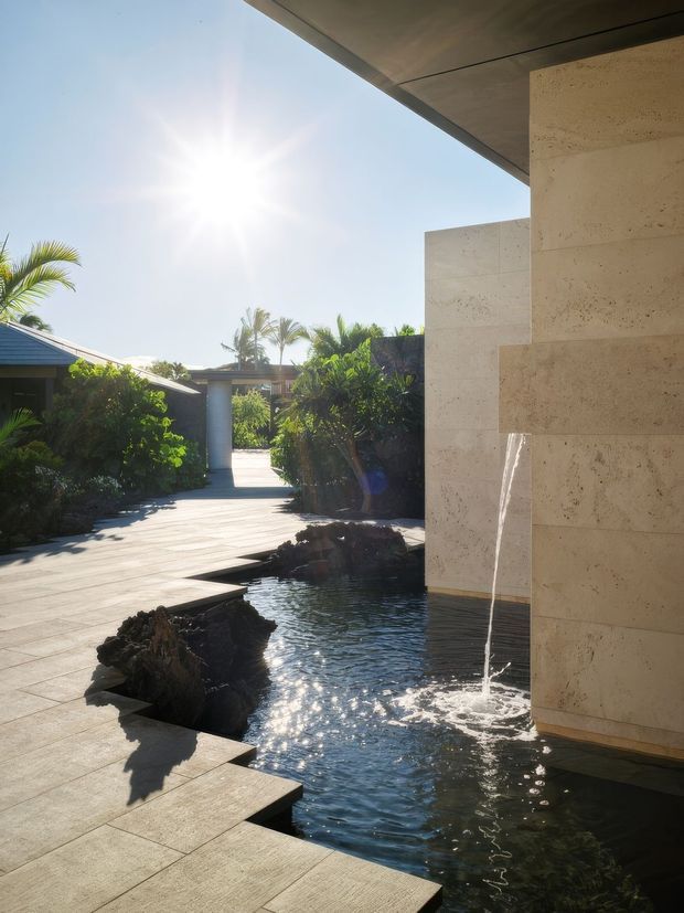 Sunlit landscaped courtyard with a shallow ornamental water feature and fountain beside a modern stone-clad building. Large rectangular paving slabs create a pathway around dark natural rocks and reflective water, with tropical planting and low-rise structures beyond. The architecture features pale textured stone walls and a broad flat projecting roof soffit.
