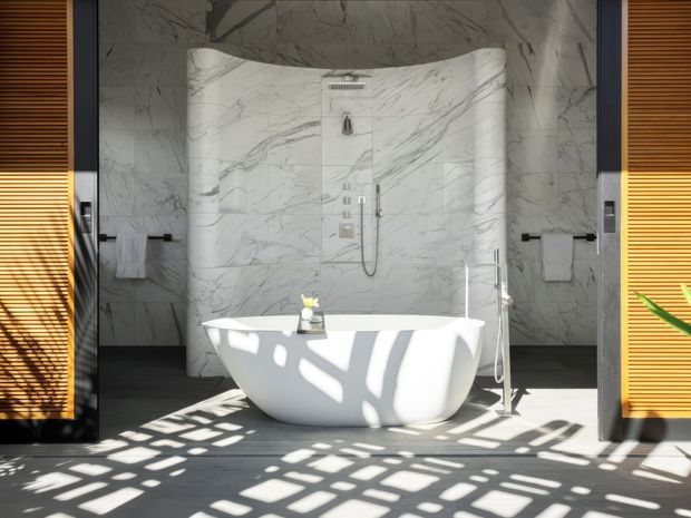 A luxurious contemporary bathroom centered on a freestanding white bathtub positioned in front of a sculptural curved marble shower wall. Large-format grey-veined marble covers the walls, while warm timber louvred screens flank the space and cast dramatic geometric shadows across the tub and floor. Chrome fixtures and simple white towels complete the refined design.
