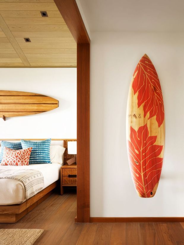 Warmly styled bedroom with a timber bed, white bedding, patterned blue and coral cushions, wooden bedside table, and a surfboard mounted as wall art. The room features rich timber flooring, a timber ceiling with recessed lighting, and a prominent dark timber structural frame or divider. Bright white walls and natural wood create a relaxed coastal atmosphere.