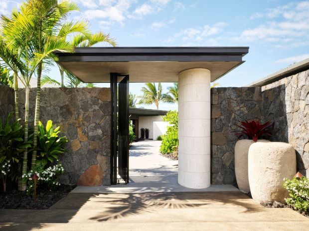 A tropical residential entry courtyard viewed from outside, with a flat-roofed portal canopy supported by a large cylindrical rendered column. Dark irregular stone walls frame a black vertical-slat gate and a paved path leading into the property. Lush palm and tropical planting, sculptural pale stone planters, and strong sunlight create a resort-like entrance.