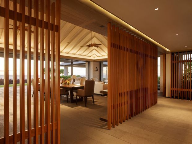 Warm, refined open-plan interior featuring extensive vertical timber screening, a timber-lined vaulted ceiling, recessed linear lighting, and a dining area opening toward expansive glazing and an outdoor view. The space uses natural wood tones, neutral furnishings, and stone or tile flooring to create a contemporary resort-like atmosphere.
