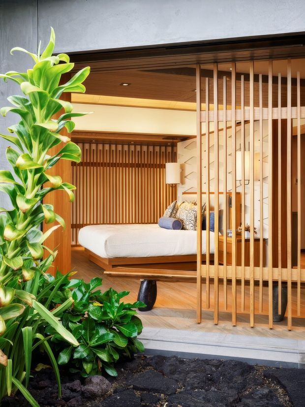 A warm, contemporary bedroom viewed from an outdoor planted entry area. The room features a low platform bed with white bedding, extensive vertical timber slat screening and wall panelling, a timber-lined ceiling, recessed lighting, and a partially open timber-framed facade. Lush tropical planting fills the foreground, creating a strong indoor-outdoor connection.
