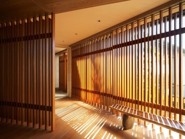 Warm, sunlit interior passage or entry space screened by extensive vertical timber battens. The timber slats create strong linear shadows across the timber floor, with a built-in or fixed timber bench along the glazed outer wall. The space has a refined contemporary architectural character with abundant natural light and strong indoor–outdoor visual connection.