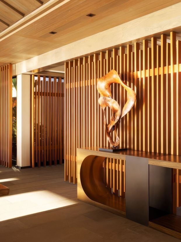 A warm, refined interior entry or hallway featuring extensive timber detailing. Vertical timber battens form a feature screen and conceal a passageway, while a timber-lined ceiling and large exposed beam span the space. A sculptural artwork sits on a built-in console with dark cabinetry and a curved timber detail. Natural light enters from the side, highlighting the rich wood tones and tiled floor.