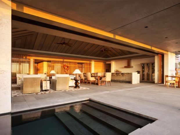 A spacious covered indoor-outdoor entertaining area opening directly onto a dark reflecting pool. The space features a timber-lined pitched ceiling with exposed beams and ceiling fans, warm integrated lighting, timber screening, lounge seating, a dining table, bar cabinetry, and large-format grey floor tiles. The overall design is luxurious, warm, and resort-like.