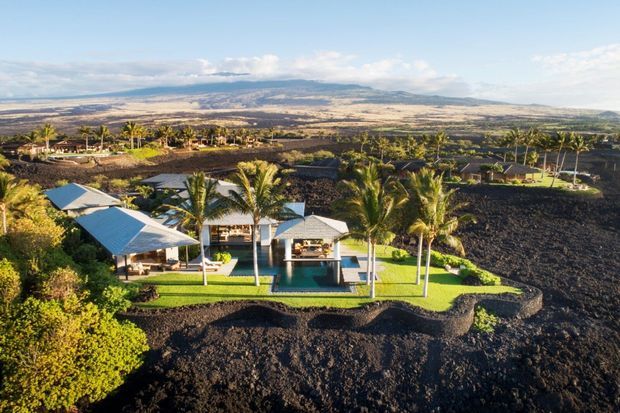 Aerial view of a tropical residential property set within a stark black volcanic landscape. The complex includes several low-profile buildings with pale metal roofs, a central open pavilion, a swimming pool, manicured lawns, and tall palm trees, with expansive dry terrain and distant mountains beyond.