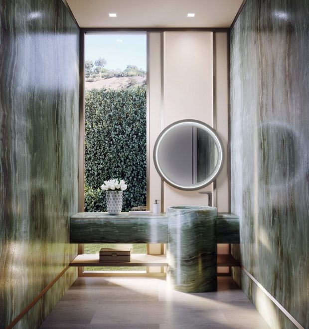 A luxurious contemporary bathroom with a sculptural cylindrical pedestal basin and matching cantilevered vanity in richly veined green stone. The room features glossy vertically textured green wall surfaces, pale panelled walls, a large round illuminated mirror, recessed ceiling lights, and a full-height window overlooking dense planting. A narrow stepped platform and warm timber detailing add architectural interest.