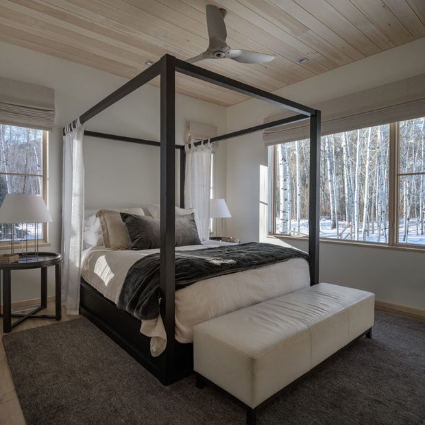 A bright, refined bedroom with a dark four-poster canopy bed, neutral bedding, a white upholstered bench, bedside table and lamp, and large windows overlooking a snowy birch forest. The room features a warm timber-lined ceiling, pale walls, and dark carpet, creating a calm contemporary retreat.