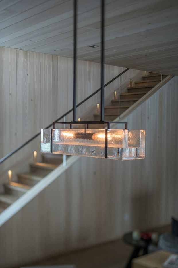 Interior architectural photograph of a refined stairway with pale vertical timber wall panelling and matching timber ceiling boards. A floating or open-riser timber staircase runs diagonally through the space, bordered by slim dark metal balustrades and illuminated by small integrated step lights. A large rectangular sculptural pendant with translucent textured glass and warm illumination hangs in the foreground. Part of a darker living area is visible below.