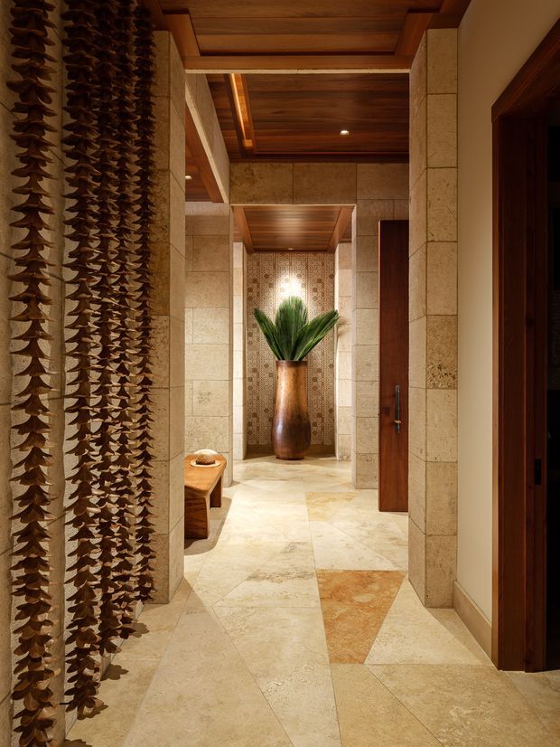 A warmly lit, richly detailed interior hallway or entry passage with cream and beige stone-clad walls and flooring, dark timber door frames, and a recessed timber-paneled ceiling. A sculptural hanging screen of layered organic forms lines the left side, while a tall wooden vessel with green foliage stands at the end against a decorative mosaic tile feature wall. The space has a refined, resort-like atmosphere with strong natural materials and geometric stone floor inlays.