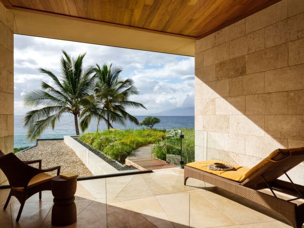 A luxurious open-air coastal terrace overlooking a tropical beach and ocean. The space features large-format beige stone wall cladding and flooring, a warm timber-lined ceiling, frameless glass balustrading, a timber deck path, and woven lounge furniture. Palm trees, ocean views, and a planted roof garden create a relaxed resort-like setting.