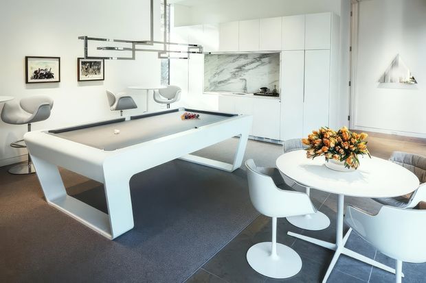 Bright, open-plan contemporary interior combining a dining area, lounge seating and a compact games or billiards table. The space features white built-in cabinetry, a marble-look splashback or feature wall, dark flooring, sculptural white chairs, a round dining table with floral arrangement, and linear pendant lighting.
