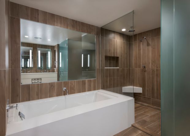 Contemporary bathroom with a large white built-in bathtub, double vanity visible in the mirror, and an open glass-enclosed shower. Warm brown vertical timber-look wall and floor tiles create a cohesive feature finish, while large mirrors and glass panels make the room feel spacious and refined.