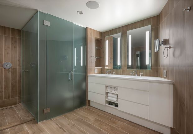 Contemporary bathroom with a large frameless glass shower enclosure, warm wood-look wall and floor tiles, and a long white double vanity. Three vertically illuminated mirrors sit above the vanity, with chrome taps and shower fittings. The room has a clean, refined appearance with recessed ceiling lighting.
